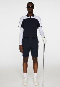 Mörkblå golfpolo med vita detaljer, parat med mörkblå shorts. Tillbehören inkluderar en vit handske, solglasögon och en golfklubba. Vita skor fulländar outfiten.