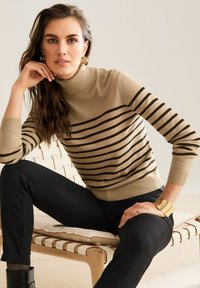 Frau mit langen lockigen Haaren, die goldene Ohrringe, einen beigen gestreiften Rollkragenpullover, schwarze Hosen und ein goldenes Armband trägt, sitzt auf einem modernen Holzstuhl.
