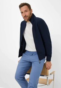 Marineblauwe zip-up jack met een hoge kraag, gedragen over een wit T-shirt, gecombineerd met lichtblauwe broek. Gladde stof en een getailleerd ontwerp.