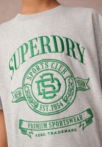 Grå bomullströja med grön broderad logotyp som har texten "Superdry", "Sports Club" och "Premium Sportswear", rund halsringning.