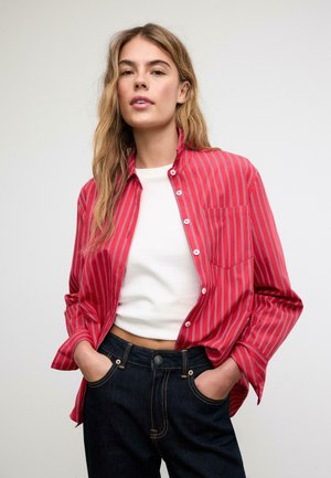 Kvinde med langt blondt hår iført en rødstribet knappet skjorte over en hvid cropped top og mørke denimjeans med hænderne i lommerne.