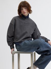 Γκρι φούτερ με ριγωτή υφή, σε oversized εφαρμογή και ψηλό λαιμό. Συνδυάζεται με μπλε τζιν παντελόνι. Κά坐 σε μεταλλική σκαμνί απέναντι από άσπρο φόντο.