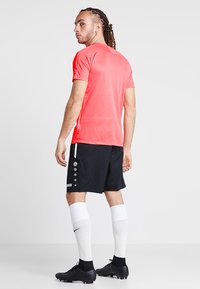 Rotes Sportshirt, schwarze Shorts mit Knopfdetails, weiße Socken und schwarze Fußballschuhe. Das Outfit hat ein körperbetontes Design für Sportbekleidung.