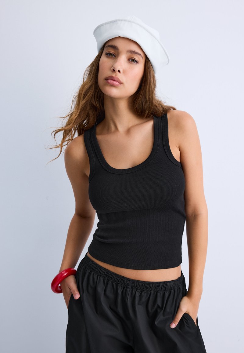 Jeune femme portant un béret blanc, un débardeur noir, un pantalon noir et un gros bracelet rouge, posant les mains dans les poches devant un fond uni.