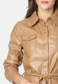 Chaqueta de camisa de cuero sintético en tono beige con cuello, dos bolsillos en el pecho, botones a presión y detalle de cintura atada. Textura suave.
