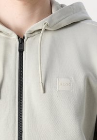 Gros plan d'un sweat à capuche zippé gris clair avec fermeture éclair noire, capuche à cordon et un petit écusson logo "BOSS" sur la poitrine.