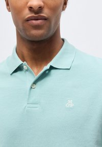 Muž nosiaci svetlomodrú polokošeľu s krátkym golierom a dvoma gombíkmi, na hrudi s malými bielymi vyšívanými písmenami "M O P".