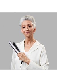 Piastra per capelli con finitura argentata e nera, tenuta da una donna in camicia bianca. Superficie liscia e design elegante. Sfondo grigio.