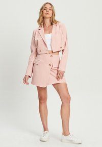 Roza skrajni blazer in krilo, ki vključuje zlate gumbe, dva sprednja žepa in gladko teksturo, v kombinaciji z belimi supergami.
