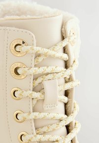 Lichtbeige veterschoen met een pluizige binnenkant, gevlochten veters met gouden accenten, gouden oogjes en een soepele leren buitenkant.