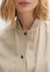 Beige jacket with a gathered neckline, featuring a smooth texture and a decorative black button closure at the collar.