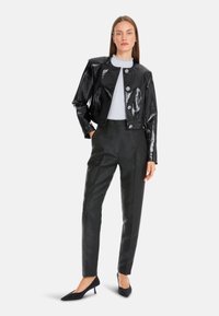 Schwarze lackierte, kurz geschnittene Jacke mit runden Knöpfen, kombiniert mit dunkel strukturierten Hosen und spitzen schwarzen Schuhen. Das Model posiert neutral.