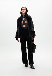 Black faux-fur jacket, patterned blouse with gold circles, black velvet trousers, accessorized with a quilted black bag and heeled boots.