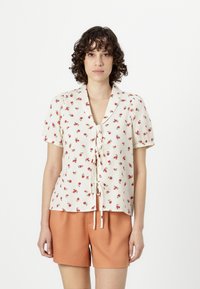 Kortermet bluse i krem med rød blomsterdesign, knyting foran, kragehals og teksturert stoff. Kombinert med oransje shorts.