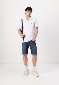Weißes Baumwoll-Polohemd mit einem gelben Schmetterlings-Logo, kombiniert mit hochgekrempelten Jeansshorts und weißen Sneakern. Glatte Textur und lässige Passform.
