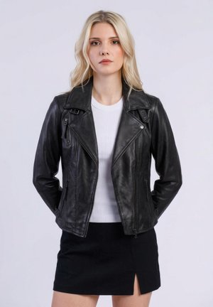 Lyshåret kvinde iført sort lædermotorcykeljakke, hvid ribbet top og sort nederdel med frontslids, stående foran en ensartet lys baggrund.