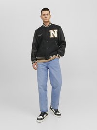 Veste de varsity noire avec des accents beiges et la lettre "N", assortie avec un jean bleu clair et des baskets noires. Tissu lisse et poignets côtelés.