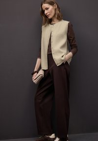 Beige geripptes Weste mit Knöpfen, getragen über einem braunen Langarmshirt, kombiniert mit braunen weitem Hosenbein und braunen Sneakers. Geflochtener Clutch in der Hand.