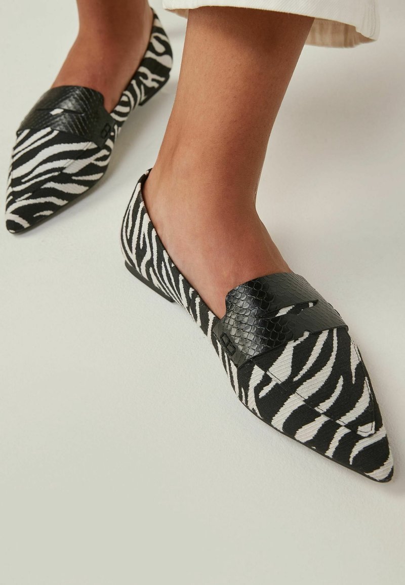 Pieds chaussés de mocassins à bout pointu imprimé zèbre noir et blanc avec des détails de bride texturée noire, sur un sol clair.