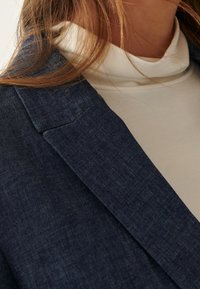 Tmavě modrý texturovaný blazer s klopami, přetažený přes světle béžový rolák. Materiál vypadá měkce a ležérně, vhodný pro různé příležitosti.