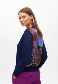Navy sweater with a floral-patterned multicolor back panel, ribbed cuffs, and waistband, worn with purple pants. Smooth texture, crew neck.