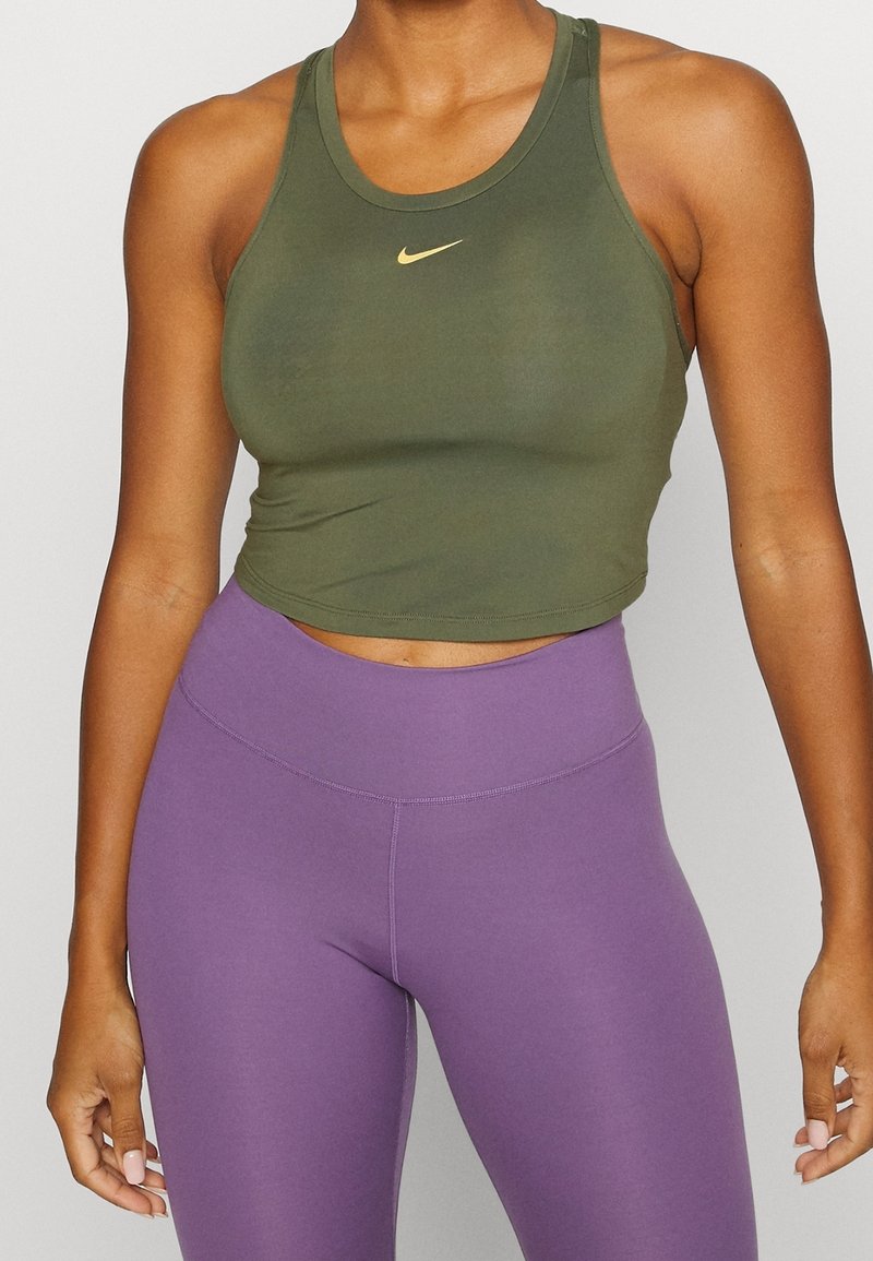 Žena v olivově zeleném bezrukávovém crop topu se žlutým logem Nike a fialovými legínami s vysokým pasem, stojící na jednobarevném pozadí.