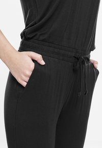 Personne portant un pantalon noir à taille élastique avec cordon, mains dans les poches avant, sur un fond clair uni.