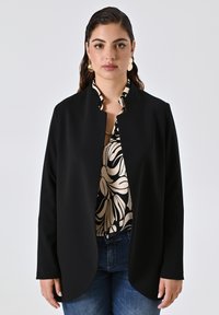 Schwarze Langarmjacke mit offener Vorderseite und abgerundetem Saum, aus glattem Stoff, über einer gemusterten Bluse mit cremefarbenen und schwarzen Designs getragen.