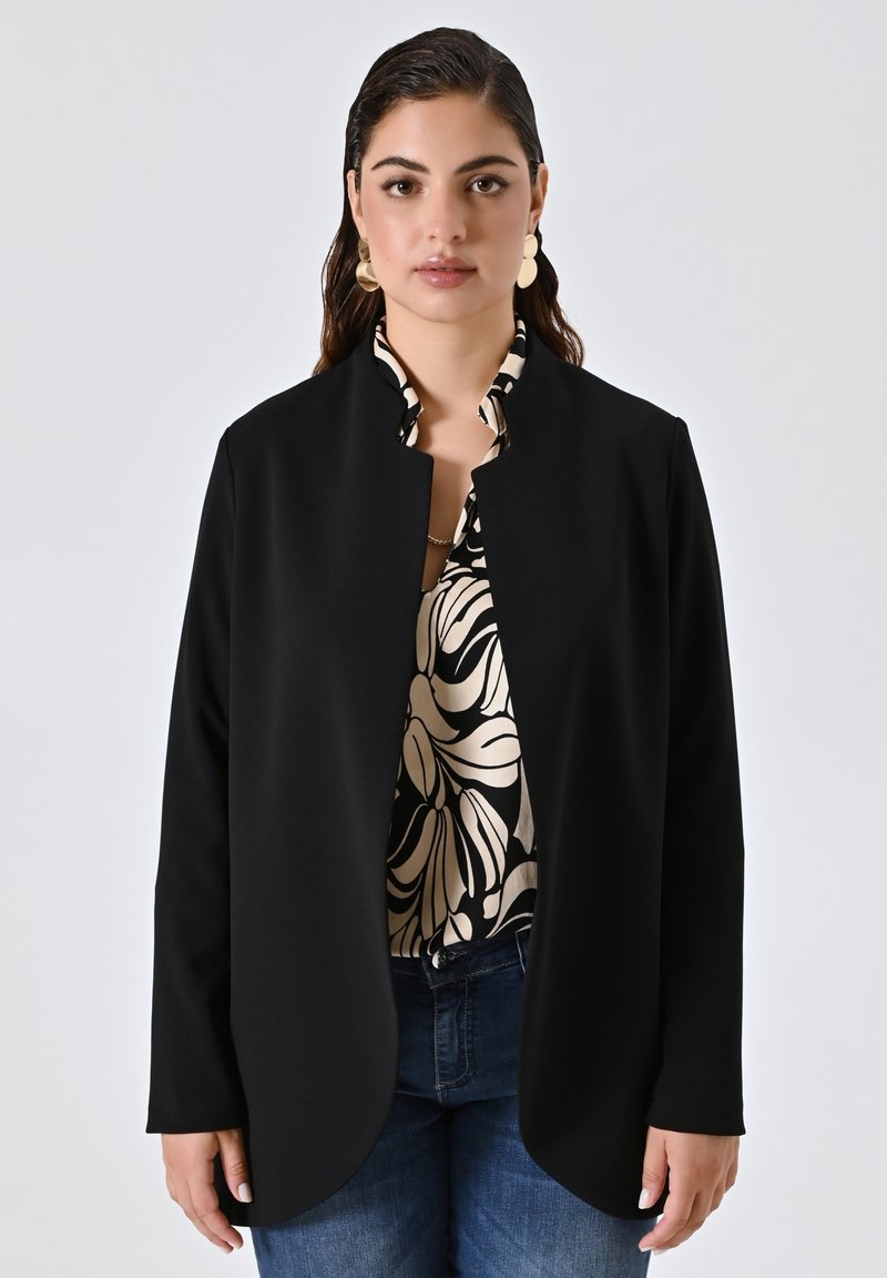 Schwarze Langarmjacke mit offener Vorderseite und abgerundetem Saum, aus glattem Stoff, über einer gemusterten Bluse mit cremefarbenen und schwarzen Designs getragen.