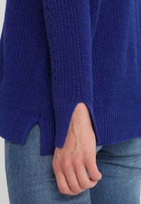  pletený svetr v jasně modré barvě, s pletenými manžetami, bočními rozparky a texturovaným vzorem, nošený přes džíny, s detailním záběrem na rukáv.