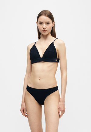Junge Frau, die ein schwarzes, strukturiertes Bikini-Set trägt, steht vor einem schlichten hellen Hintergrund und blickt mit neutralem Gesichtsausdruck nach vorne.