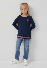 Marineblaues Langarmoberteil mit einem pinken Herzdetail, überlagert mit einem floralen pinken Volantsaum, kombiniert mit hellblauen Jeans und weißen Turnschuhen.