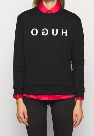 Personne portant un sweat-shirt noir avec le texte blanc inversé "HUGO" sur une chemise rouge à col, associé à un pantalon noir, sur un fond uni.