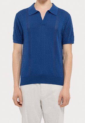 Mann trägt ein blaues kurzärmeliges Strick-Poloshirt mit strukturierten vertikalen Streifen und weiße Hose vor einfarbigem Hintergrund.