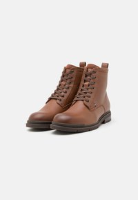 Bottes chelsea en cuir marron avec une surface lisse et texturée, bout arrondi, lacets à l'avant et un logo discret sur la semelle.