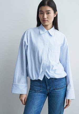 Mladá žena v nadrozmernej svetlomodro-bielo pruhovanej košeli s širokými manžetami a vysoko pásovými modrými džínsami stojaca oproti jednoliatej stene.