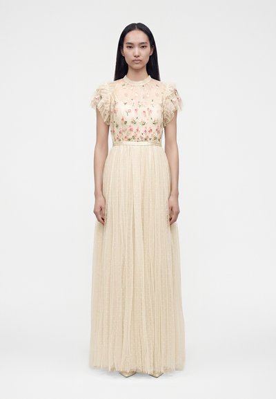 Femme portant une longue robe en tulle beige avec une broderie florale sur le corsage et des manches courtes volantées, debout devant un fond uni.