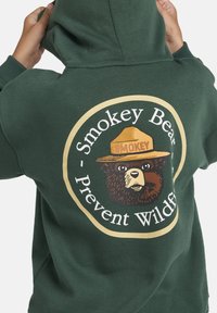 Grüne Kapuzenjacke mit einem Grafikmotiv von Smokey Bear, das einen braunen Bären mit Hut zeigt. Der Text lautet "Waldbrände verhindern." Weiches Baumwollmaterial.