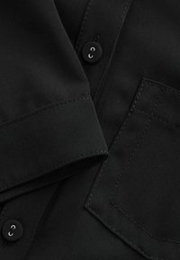 Schwarzer Stoff mit einer glatten Textur. Verfügt über einen Knopf, eine Manschette und eine aufgesetzte Tasche. Die Nähte sind sauber und gleichmäßig.