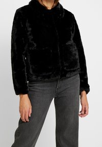 Svart fuskpälsjacka med mjuk textur, med krage, två framfickor och en kortad design, kombinerad med mörkgrå jeans.