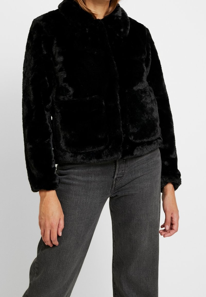 Svart fuskpälsjacka med mjuk textur, med krage, två framfickor och en kortad design, kombinerad med mörkgrå jeans.