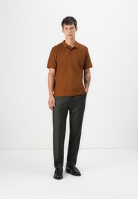 Smeđa polo majica s kratkim rukavima u kombinaciji s tamnozelenim hlačama; ima klasičnu ovratnik i suptilnu teksturu. Crne cipele upotpunjuju outfit.