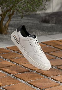 White leather sneaker with a black heel cap, flat laces, perforated accents, and a subtle logo on the side, set against textured tiles.