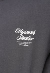 Sweatshirt cinza escuro com um logo estilizado branco que inclui o texto "Originals Studio" e detalhes adicionais da fundação abaixo.