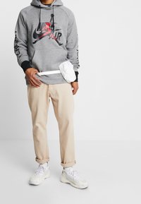 Sudadera gris con detalles negros y gráficos rojos, pantalones beige, riñonera blanca y zapatillas blancas en un atuendo casual.