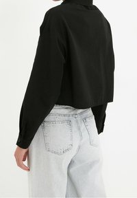 Osoba w luźnej, czarnej koszuli z długim rękawem, włożonej w jasnoniebieskie, wysokie dżinsy z widocznymi tylnymi kieszeniami, pokazana od tyłu.