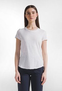 Camiseta blanca de manga corta con tejido ligero y ligeramente texturizado; cuello redondeado y dobladillo curvado. Combinada con pantalones oscuros.