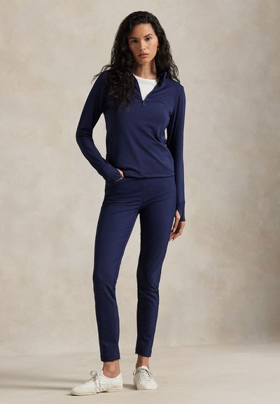 Vrouw met lang donker haar staat in een marineblauwe zip-up top, bijpassende broek, wit overhemd eronder en witte sneakers tegen een effen achtergrond.