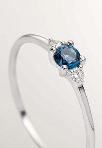 Anillo de plata con una piedra preciosa azul ovalada, flanqueada por dos piedras claras más pequeñas. Banda suave con un acabado pulido.