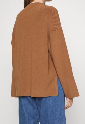 Personne portant un pull en maille marron ample avec des fentes latérales et un pantalon bleu, vue de dos sur un fond clair uni.